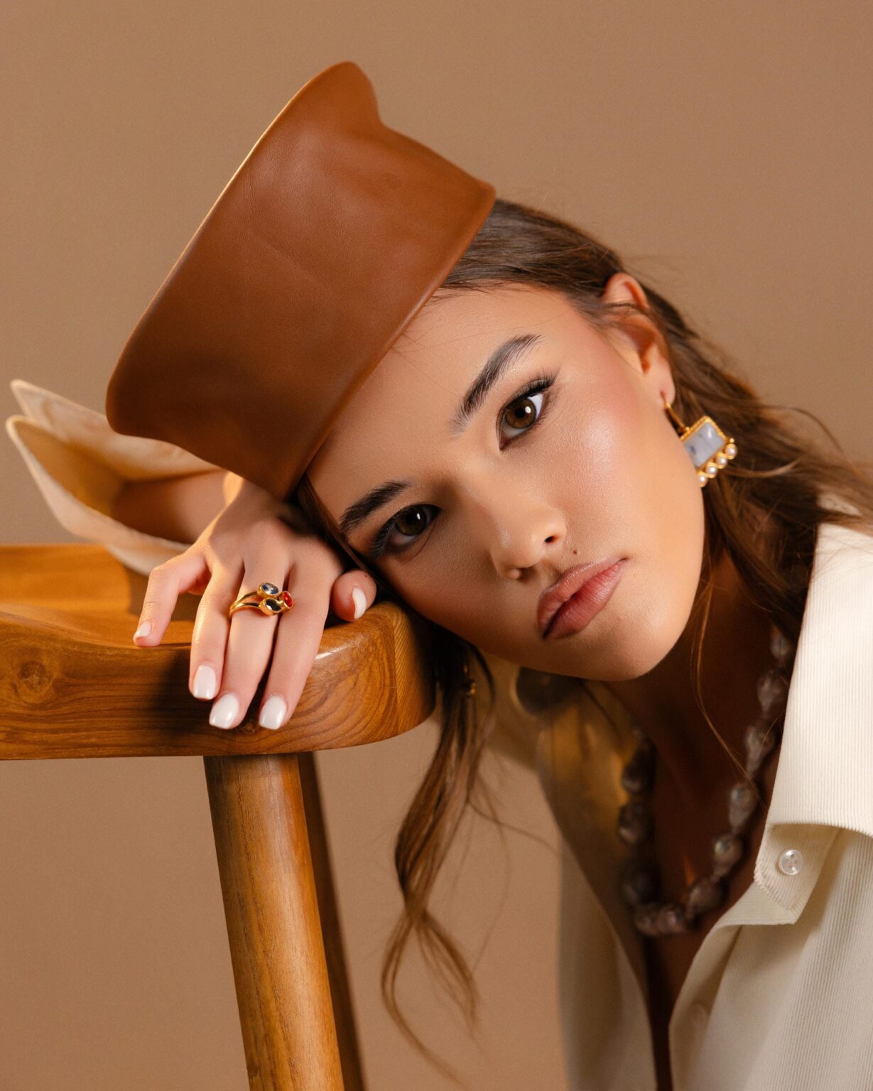Model in a tan leather hat wearing a gemstone gold ring and pearl-trimmed earrings